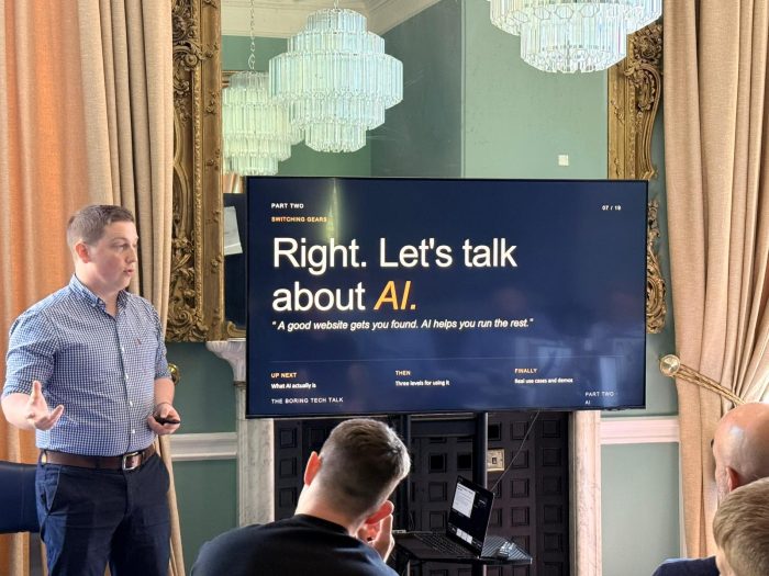 Sam presenting beside a 'Let's talk about AI' slide to a seated audience