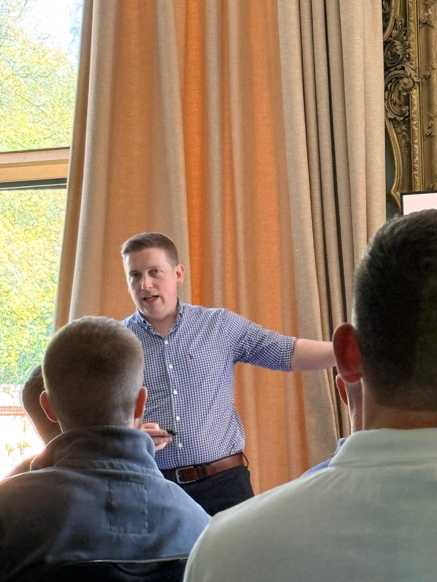 Sam reaching out towards the audience while presenting, attendees in the foreground